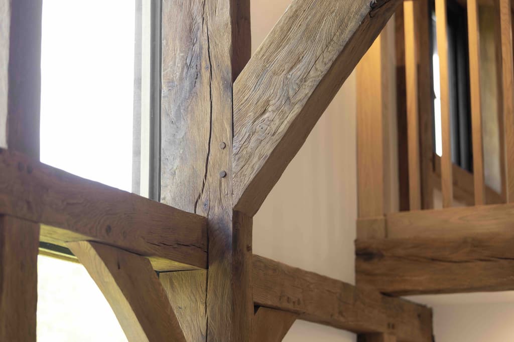 Close detail of weathered oak posts and braces joined with timber pegs, beside a tall window with an oak balustrade beyond