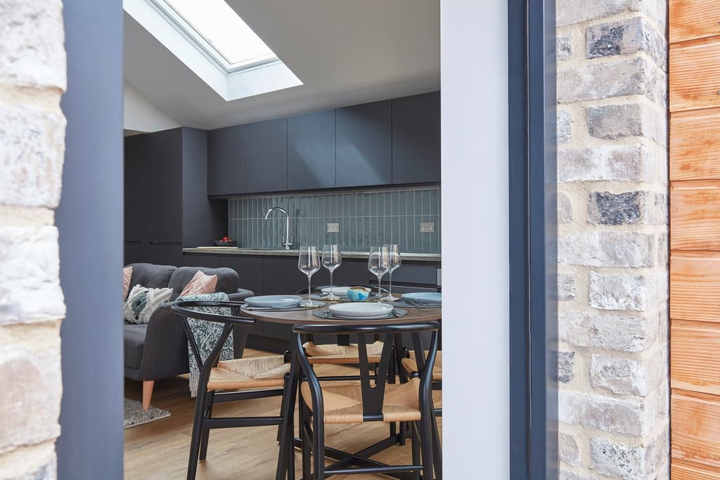 View through an open door, between pale brick and timber boarding, to the dining table and dark grey kitchen under a rooflight