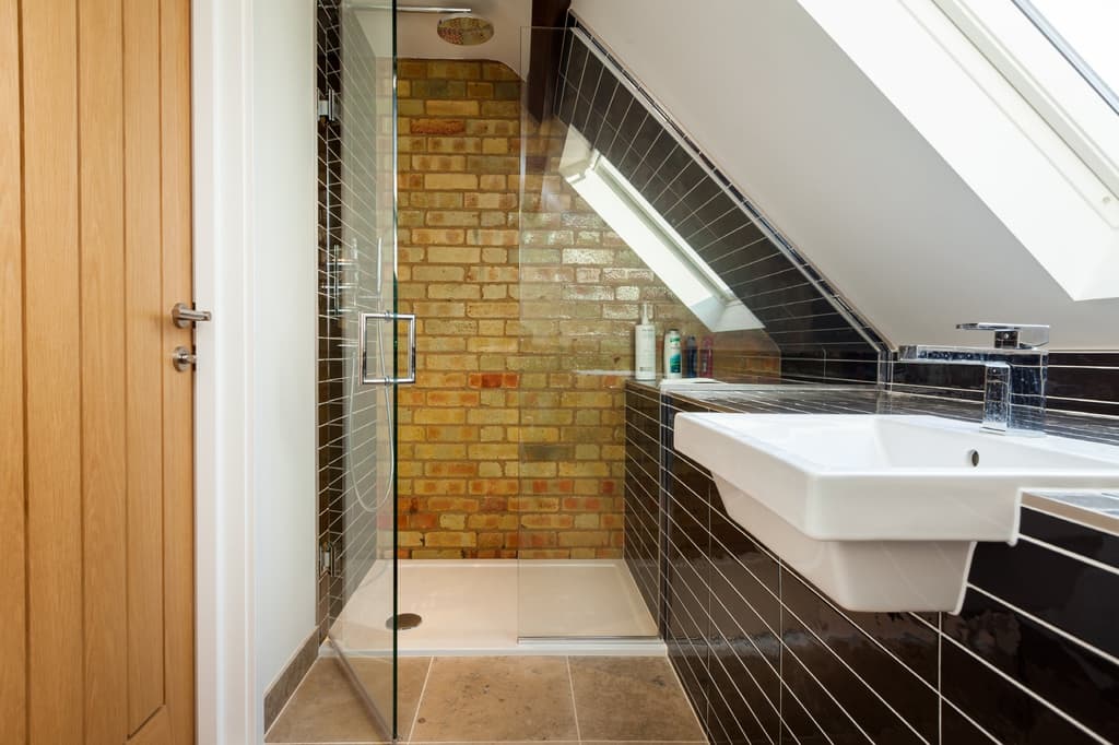 Shower room under a sloping ceiling: a frameless glass enclosure against exposed yellow brick, dark glossy wall tiles, a rooflight and stone floor tiles