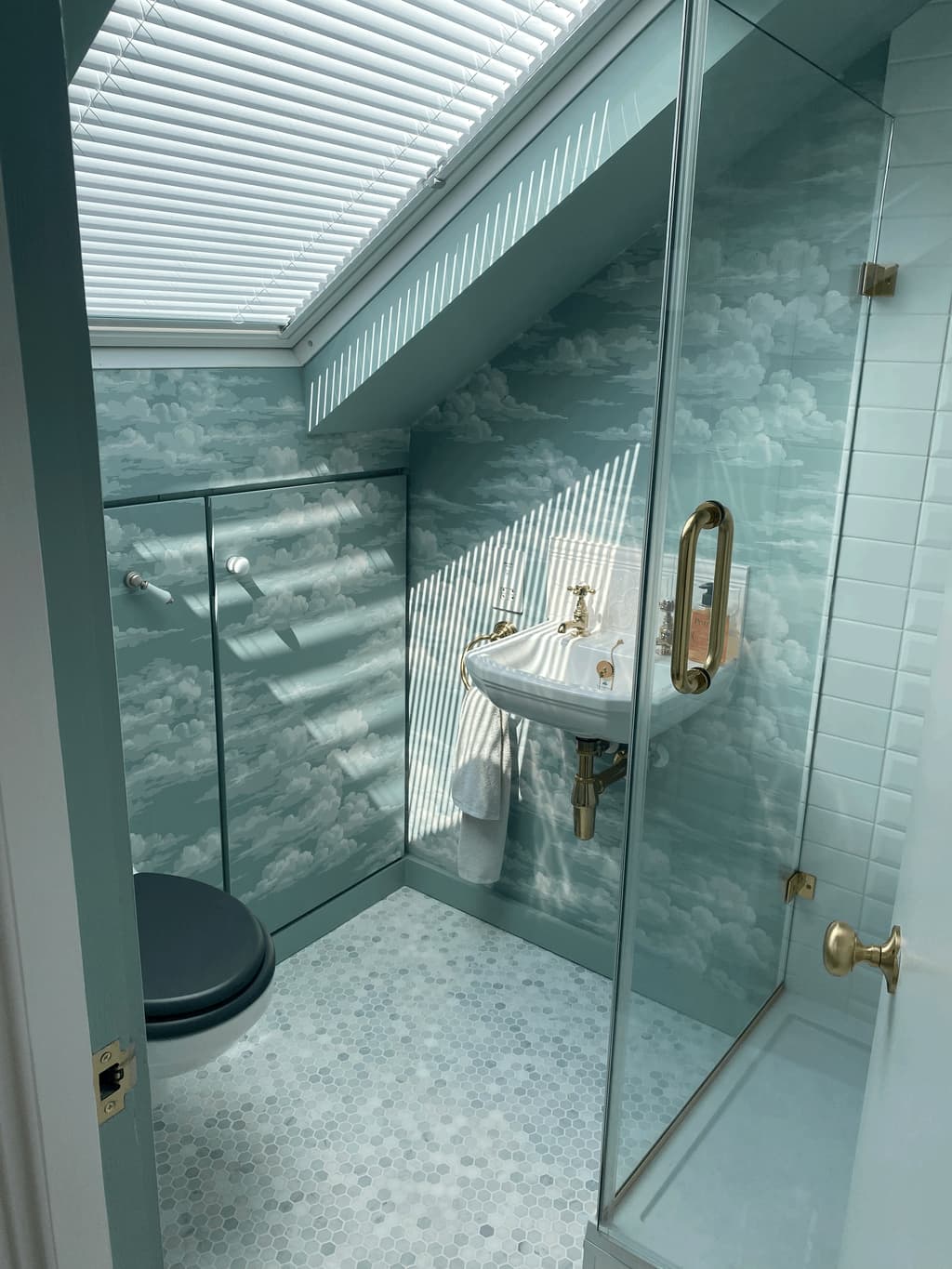 En-suite with cloud-pattern wallpaper, a hexagonal mosaic floor, a brass-fitted basin and a glass shower screen, striped with light from the rooflight blind