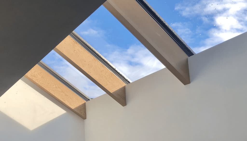 Looking up through glazed rooflights set between timber beams to a blue sky with cloud