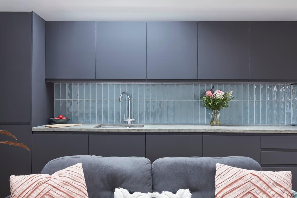 Dark grey handleless kitchen with a blue glazed vertical-tile splashback and grey worktop, seen over the back of a sofa