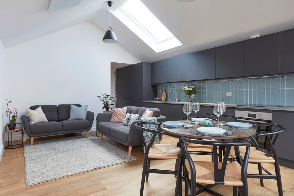 Open-plan room under a vaulted ceiling with a rooflight: two grey sofas, a round dining table and a dark grey kitchen with blue glazed tiles