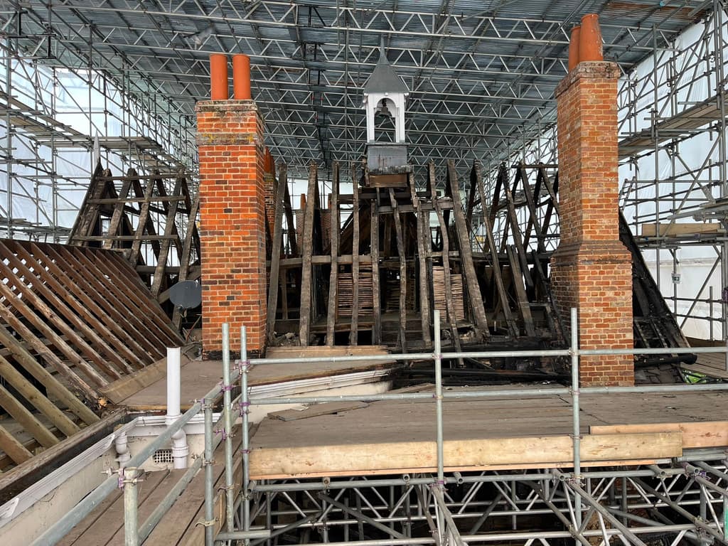 Two brick chimney stacks and the bell turret rising among exposed roof timbers inside the temporary scaffold roof during repair