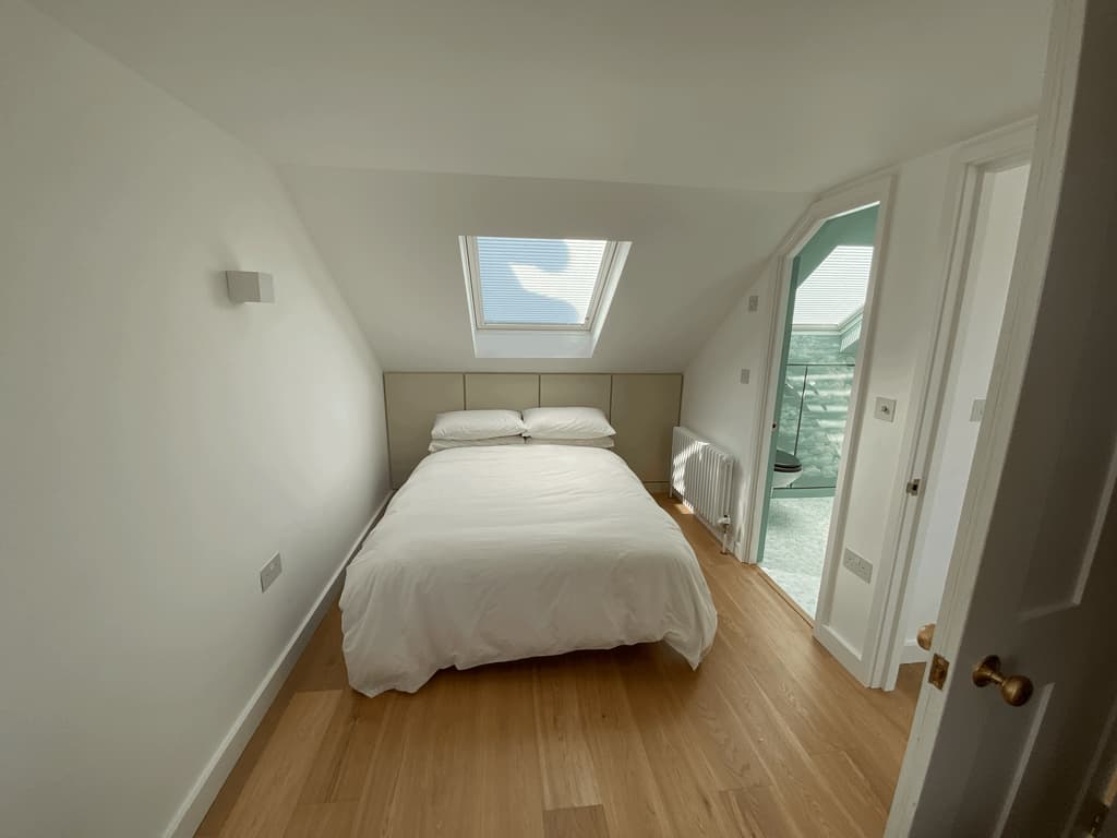 Loft bedroom under a sloping ceiling, lit by a rooflight, with oak floorboards and a door open to a teal en-suite