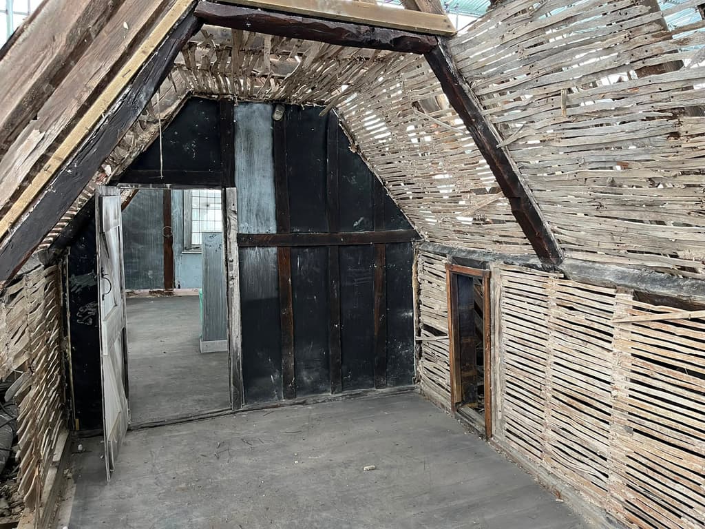 Attic room stripped to riven timber lath on walls and ceiling, with an exposed timber frame and a dark boarded partition with a doorway