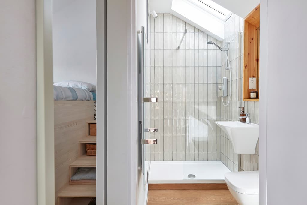 Shower room with white glazed vertical tiles, a rooflight and a wall-hung basin, seen from the bedroom beside the raised bed's steps