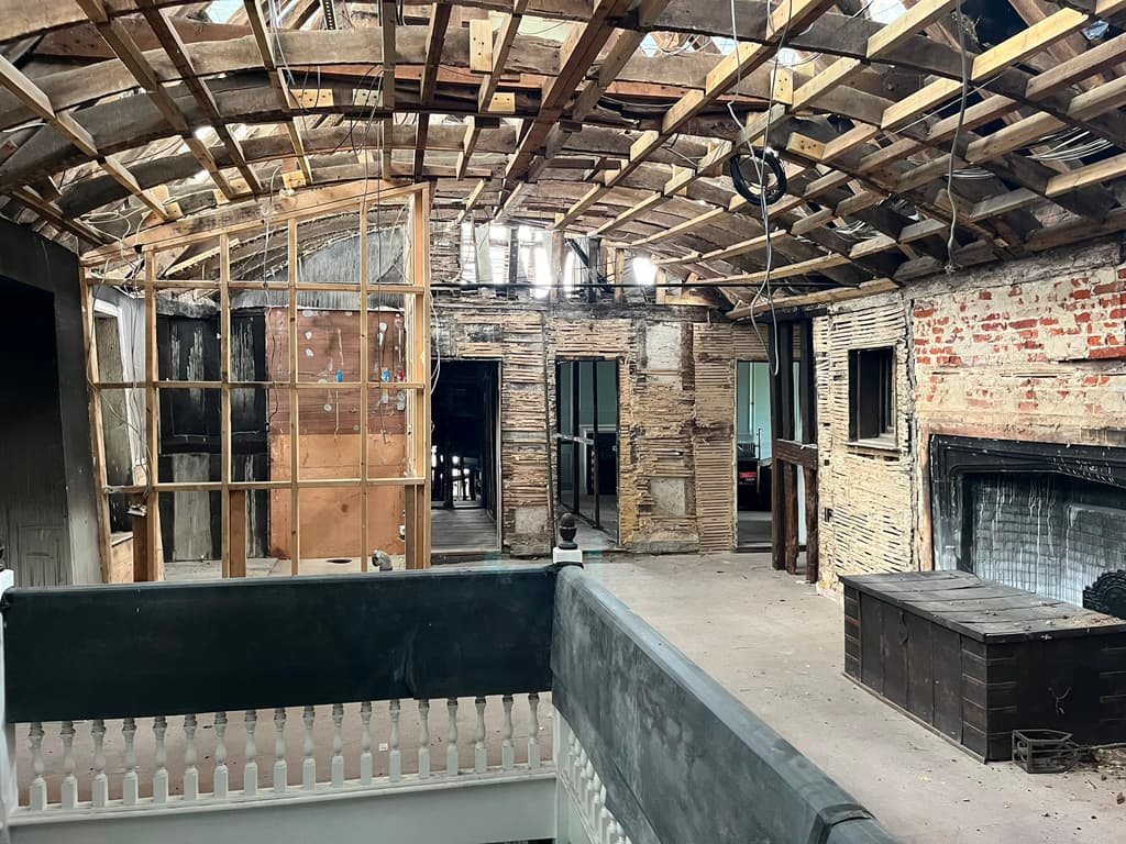 Upper floor stripped back during repair: exposed lath and brick walls, new pale roof timbers among the old, a stair balustrade and a large fireplace