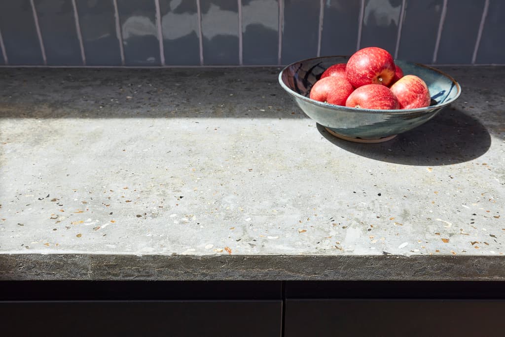 Sunlit speckled grey worktop with a ceramic bowl of red apples against blue glazed tiles and dark cabinet fronts
