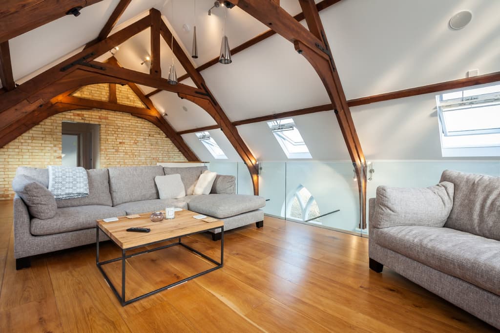 Gallery sitting room beneath arched timber roof trusses, with a yellow-brick gable, oak floorboards, rooflights and a glass balustrade