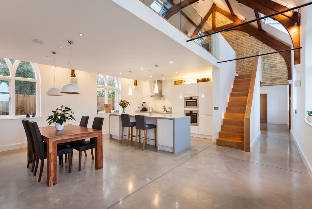 Double-height kitchen and dining space in the converted church: polished concrete floor, arched windows, an oak stair and a glass-balustraded gallery beneath exposed roof trusses