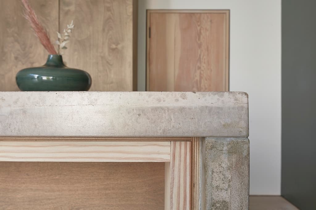 Edge of a concrete worktop on a pale timber frame, with a green ceramic vase and plywood cabinets behind