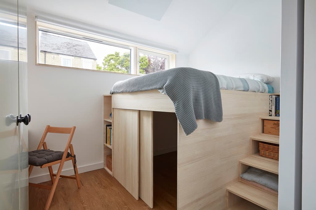 Bedroom with a pale timber raised bed over sliding-door storage, built-in storage steps and a long window towards neighbouring roofs