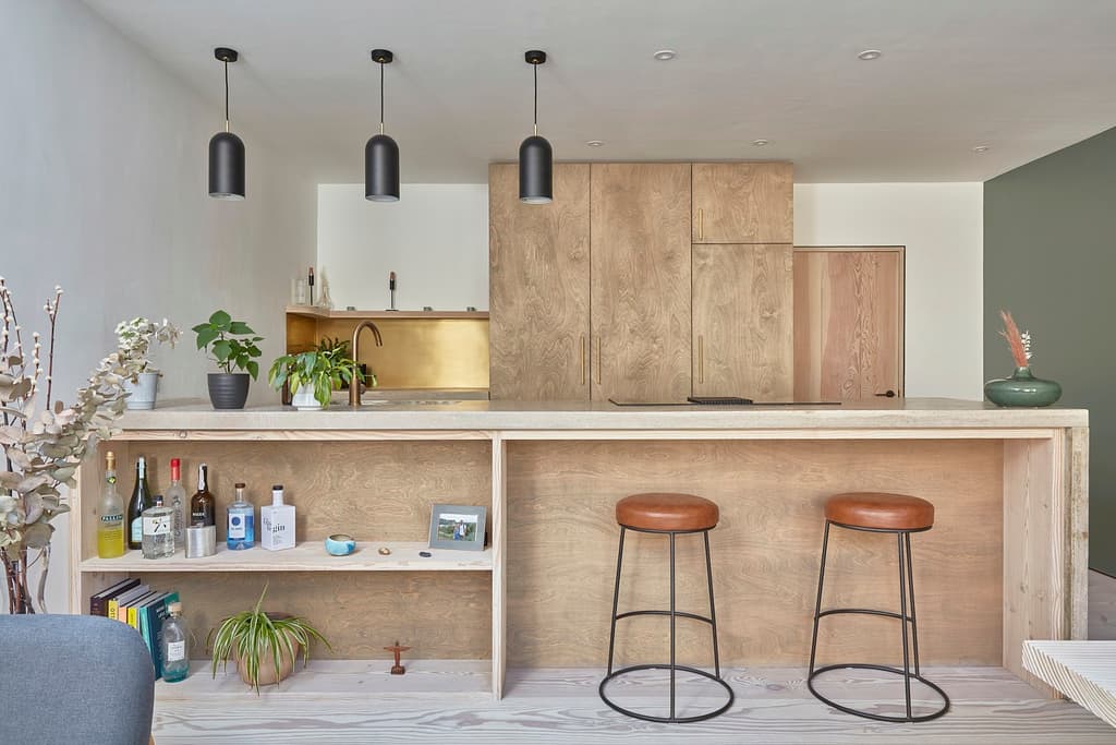 Kitchen island with a pale timber frame and concrete worktop, leather-topped stools, tall plywood cabinets and a brass splashback