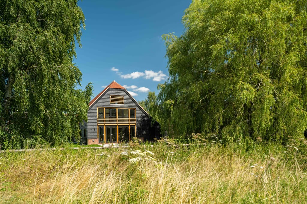 The Oak Barn's black weatherboarded gable with two storeys of oak-framed glazing under a clay-tiled half-hip, seen across a wildflower meadow between a silver birch and a weeping willow