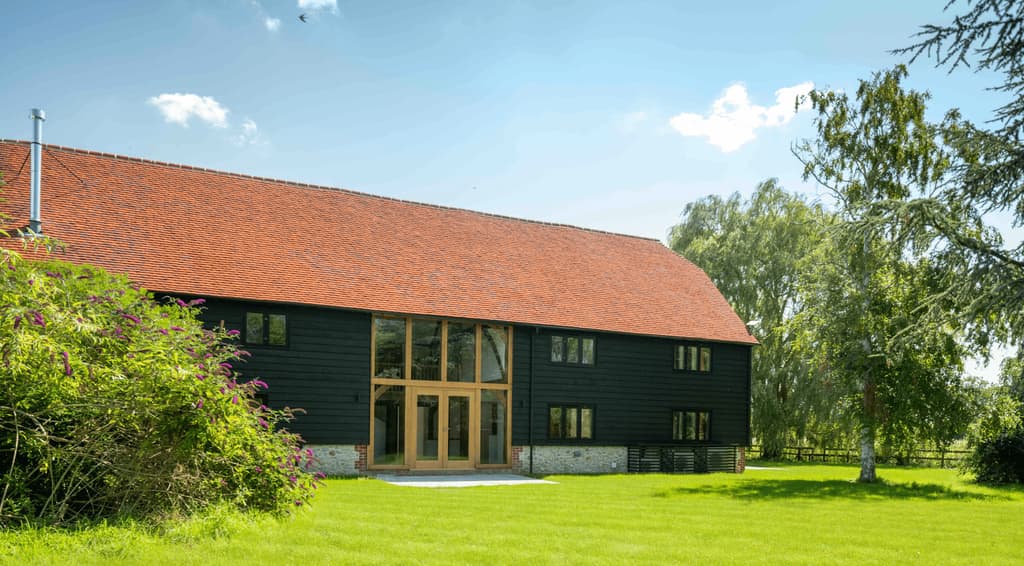 Long elevation of The Oak Barn: red clay-tiled roof, black weatherboarding on a flint plinth and a full-height oak-framed glazed entrance facing the lawn